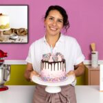 Mujer sonriente sosteniendo un pastel decorado con chocolate y chispas en una cocina con fondo rosa. A su izquierda hay una batidora roja y a su derecha un libro de recetas 'Anyone Can Cake' y cupcakes. - Recetas Chidas
