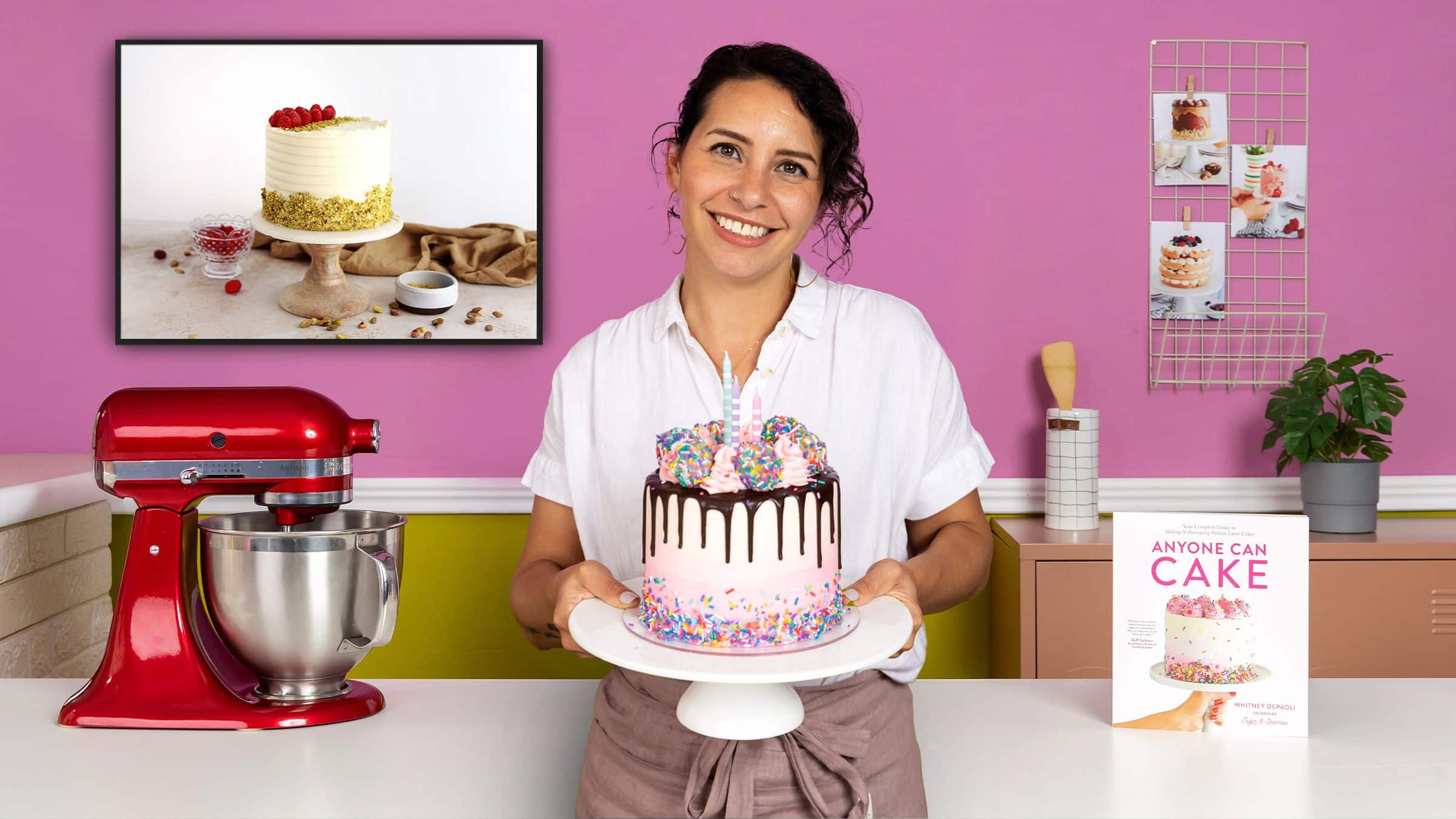 Mujer sonriente sosteniendo un pastel decorado con chocolate y chispas en una cocina con fondo rosa. A su izquierda hay una batidora roja y a su derecha un libro de recetas 'Anyone Can Cake' y cupcakes.