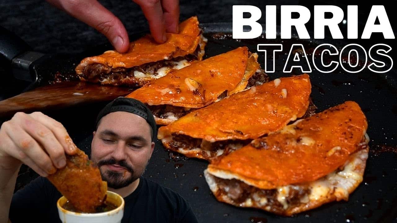Imagen de tacos de birria: varios tacos dorados rellenos de carne sobre una plancha, con una persona sumergiendo uno en un tazón de consomé y el texto "BIRRIA TACOS".
