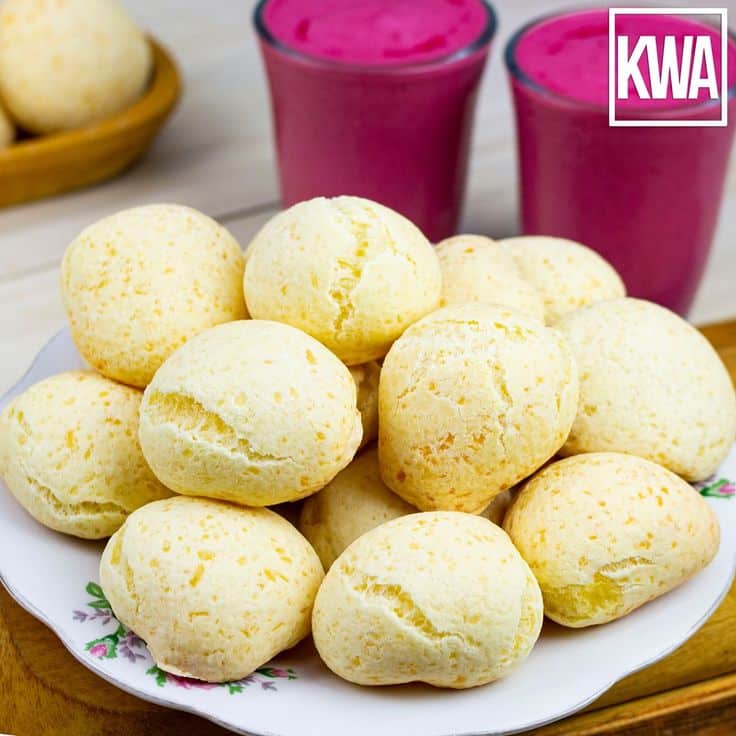 Plato de panecillos de yuca horneados en forma de bolitas amarillas sobre un plato decorado, con dos vasos de bebida rosa en el fondo y el logotipo 'KWA' en la esquina. - Recetas Chidas