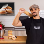 Hombre con gorra y delantal negro posando en la cocina frente a una batidora roja y una torre de donas apiladas, con un cuadro de donas en la pared de fondo. - Recetas Chidas