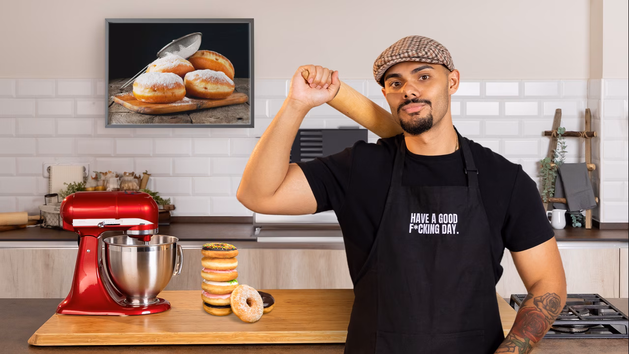 Hombre con gorra y delantal negro posando en la cocina frente a una batidora roja y una torre de donas apiladas, con un cuadro de donas en la pared de fondo. - Recetas Chidas