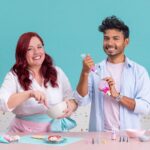 Dúo de reposteros alegres decorando galletas con royal icing en una cocina de tonos pastel. La mujer pelirroja mezcla glaseado en un bol rosa mientras el hombre sostiene una espátula y una piruleta; la mesa está llena de cupcakes y adornos. - Recetas Chidas