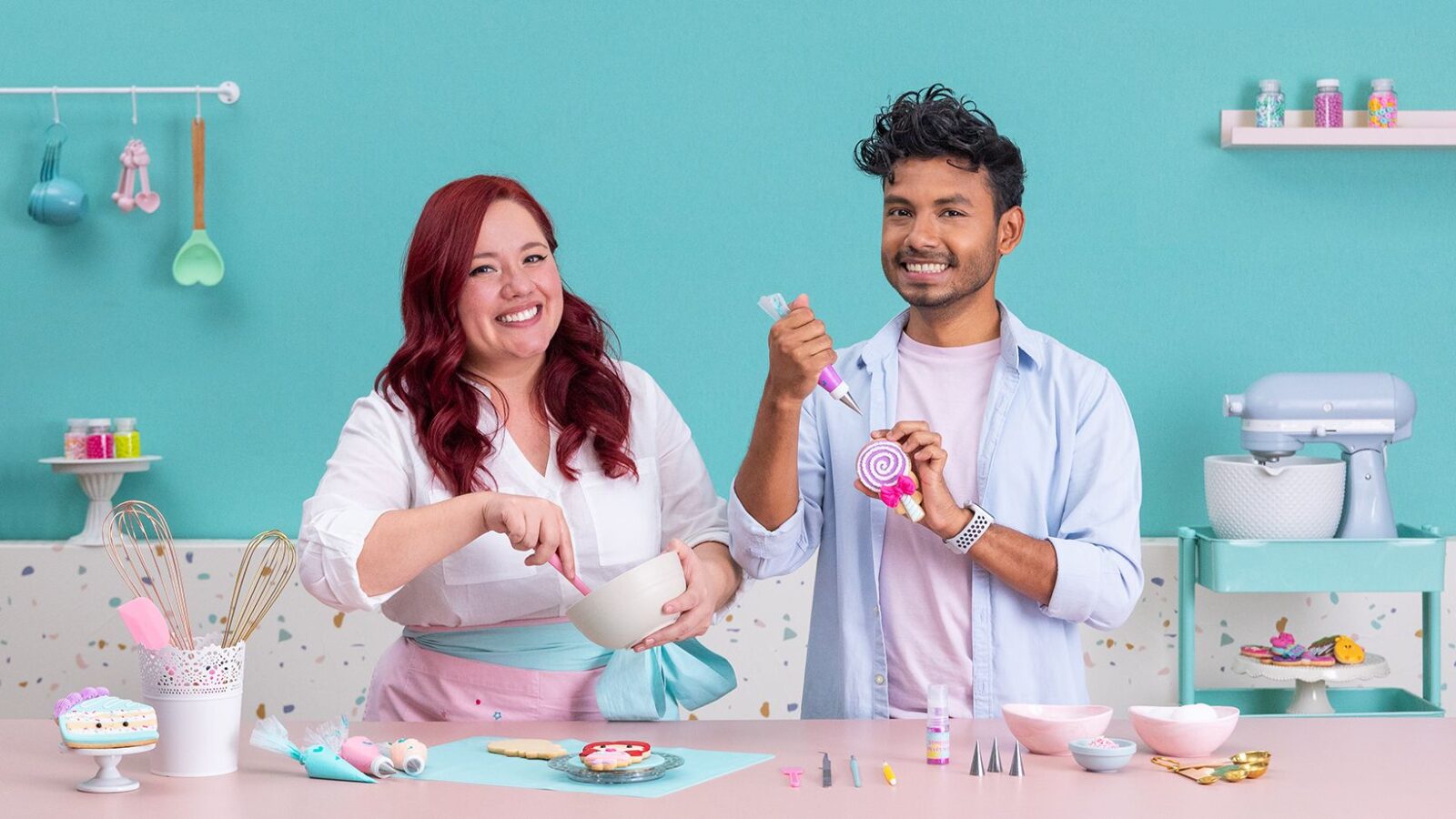 Dúo de reposteros alegres decorando galletas con royal icing en una cocina de tonos pastel. La mujer pelirroja mezcla glaseado en un bol rosa mientras el hombre sostiene una espátula y una piruleta; la mesa está llena de cupcakes y adornos. - Recetas Chidas