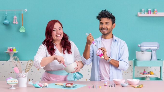 Dúo de reposteros alegres decorando galletas con royal icing en una cocina de tonos pastel. La mujer pelirroja mezcla glaseado en un bol rosa mientras el hombre sostiene una espátula y una piruleta; la mesa está llena de cupcakes y adornos. - Recetas Chidas
