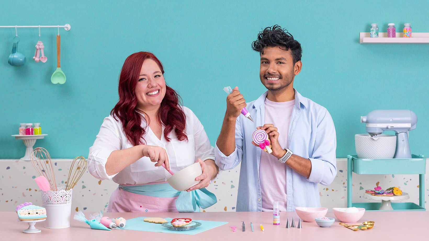 Dúo de reposteros alegres decorando galletas con royal icing en una cocina de tonos pastel. La mujer pelirroja mezcla glaseado en un bol rosa mientras el hombre sostiene una espátula y una piruleta; la mesa está llena de cupcakes y adornos.