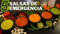Imagen de cinco salsas servidas en pequeños recipientes de piedra sobre una mesa negra, rodeadas de ingredientes como chiles verdes, tomates, ajos y un aguacate, con texto grande '5 salsas de emergencia' en verde y blanco. - Recetas Chidas