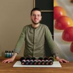 Chef joven con gafas y chaqueta de cocina presenta una bandeja con una fila de bombones de chocolate de colores brillantes, de pie en una cocina con un fondo desenfocado de bombones. - Recetas Chidas