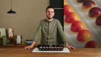 Chef joven con gafas y chaqueta de cocina presenta una bandeja con una fila de bombones de chocolate de colores brillantes, de pie en una cocina con un fondo desenfocado de bombones. - Recetas Chidas
