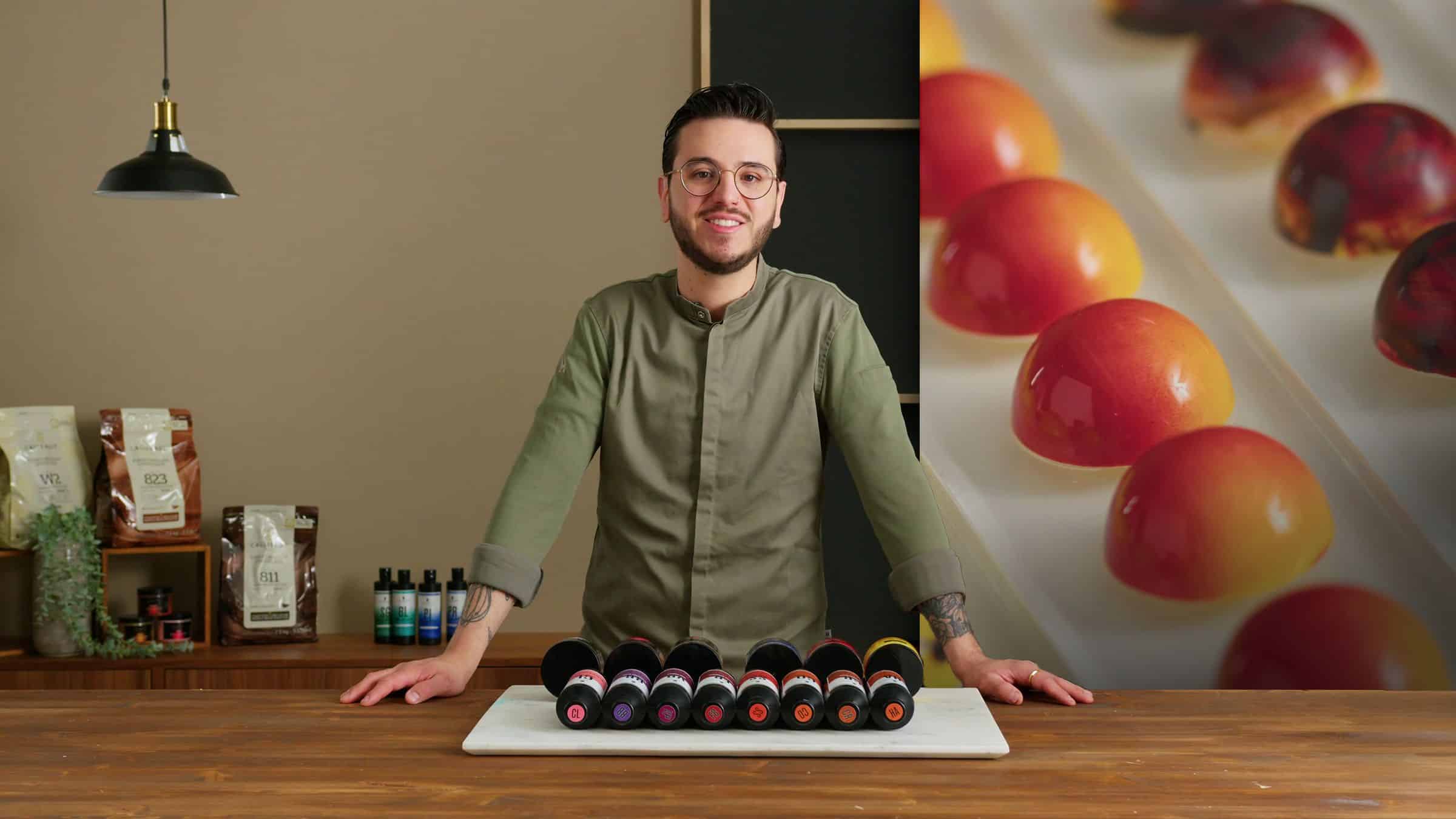 Chef joven con gafas y chaqueta de cocina presenta una bandeja con una fila de bombones de chocolate de colores brillantes, de pie en una cocina con un fondo desenfocado de bombones.