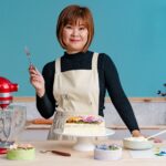 Mujer asiática con delantal blanco decorando un pastel con flores de crema de mantequilla usando una espátula, junto a una batidora roja y varios pasteles coloridos en una cocina de tonos pastel. - Recetas Chidas