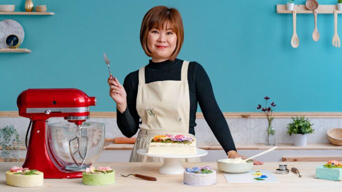 Mujer asiática con delantal blanco decorando un pastel con flores de crema de mantequilla usando una espátula, junto a una batidora roja y varios pasteles coloridos en una cocina de tonos pastel. - Recetas Chidas