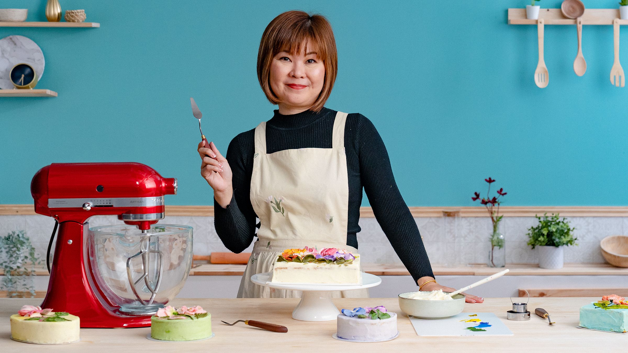 Mujer asiática con delantal blanco decorando un pastel con flores de crema de mantequilla usando una espátula, junto a una batidora roja y varios pasteles coloridos en una cocina de tonos pastel.