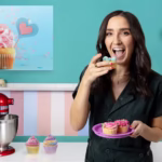 Mujer sonriente vestida de negro degustando y decorando cupcakes con glaseado colorido en una cocina, con una batidora roja y un fondo turquesa con cuadro de cupcake. - Recetas Chidas