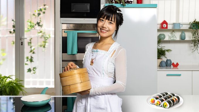 Joven mujer asiática con delantal blanco sostiene una cesta de bambú en una cocina luminosa mientras sonríe, con un plato de rollos de sushi veganos de colores sobre la mesa y una sartén turquesa a la izquierda.