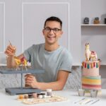 Pastelero sonriente modelando figuritas de animales de chocolate, sentado en una mesa con pinturas y herramientas. A su lado hay un pastel de jirafa y un pastel con forma de tronco decorado con flores y frutas. - Recetas Chidas