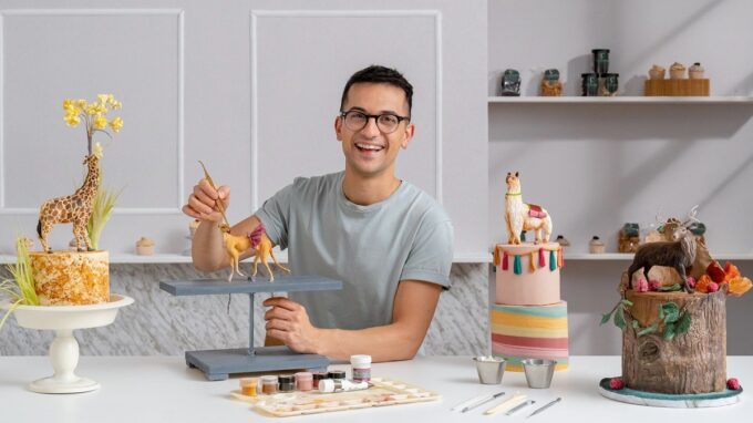 Pastelero sonriente modelando figuritas de animales de chocolate, sentado en una mesa con pinturas y herramientas. A su lado hay un pastel de jirafa y un pastel con forma de tronco decorado con flores y frutas.