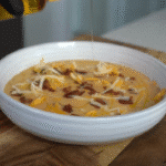 Sopa cremosa de papa y queso servida en un tazón blanco con trozos de tocino y queso rallado, mientras se vierte una salsa o queso derretido por encima. - Recetas Chidas