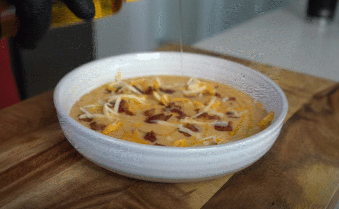 Sopa cremosa de papa y queso servida en un tazón blanco con trozos de tocino y queso rallado, mientras se vierte una salsa o queso derretido por encima.