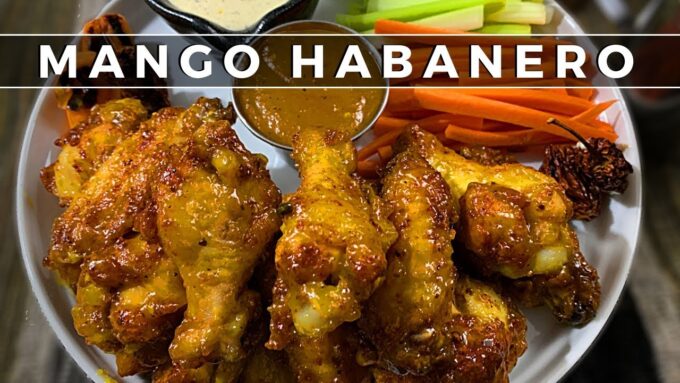 Plato de alitas de pollo cubiertas con salsa mango habanero acompañadas de bastones de zanahoria y apio y salsa dip en el centro, con texto sobrepuesto 'MANGO HABANERO'.