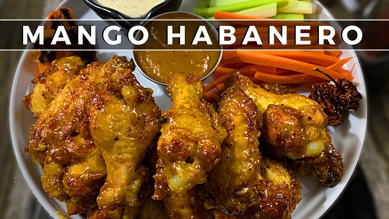 Plato de alitas de pollo cubiertas con salsa mango habanero acompañadas de bastones de zanahoria y apio y salsa dip en el centro, con texto sobrepuesto 'MANGO HABANERO'.