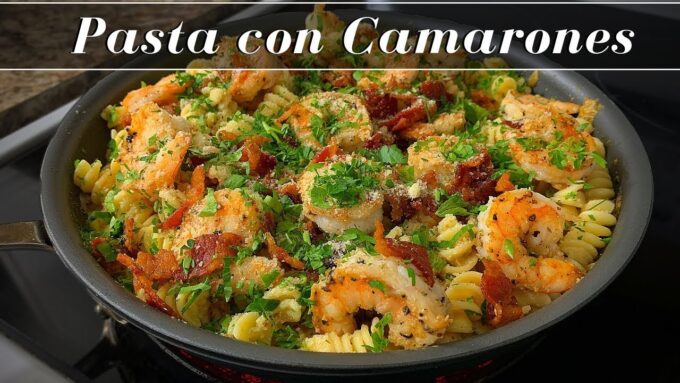 Sartén con pasta fusilli salteada con camarones, trozos de tocino y perejil. En la parte superior de la imagen aparece el texto "Pasta con Camarones".