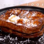 Tostadas francesas gruesas apiladas en un plato negro, con mantequilla, jarabe y azúcar glass espolvoreada; el texto "French Toast" aparece superpuesto. - Recetas Chidas