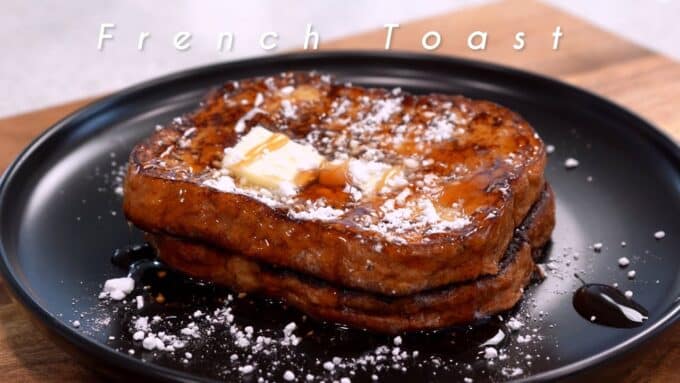Tostadas francesas gruesas apiladas en un plato negro, con mantequilla, jarabe y azúcar glass espolvoreada; el texto "French Toast" aparece superpuesto.
