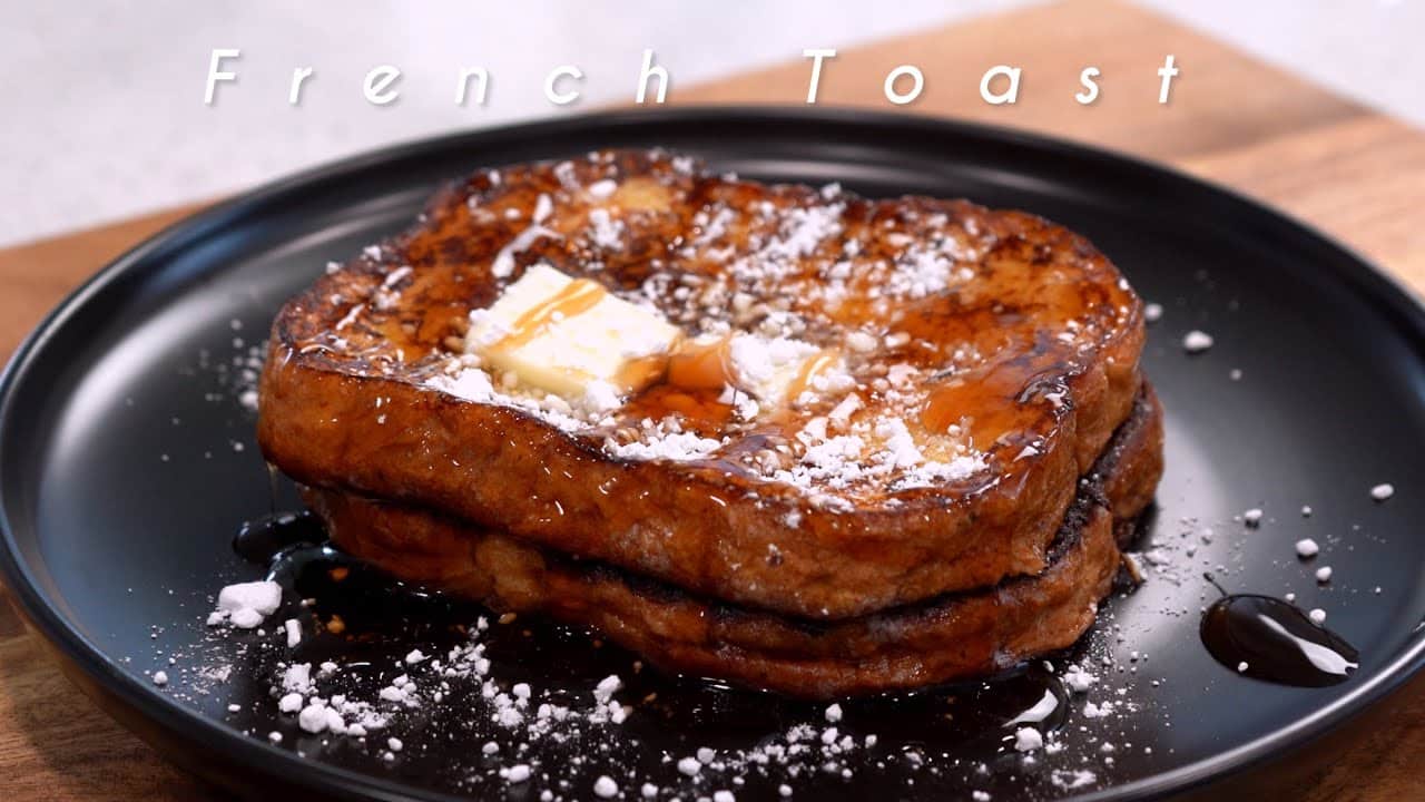 Tostadas francesas gruesas apiladas en un plato negro, con mantequilla, jarabe y azúcar glass espolvoreada; el texto "French Toast" aparece superpuesto. - Recetas Chidas