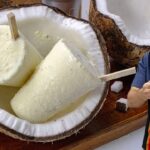 Paletas de helado de coco artesanales servidas dentro de cáscaras de coco, con un hombre sosteniendo y probando una de ellas. - Recetas Chidas