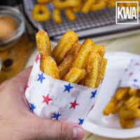 Mano sosteniendo un vaso decorado con estrellas lleno de churros fritos en primer plano, con más churros desenfocados de fondo. - Recetas Chidas